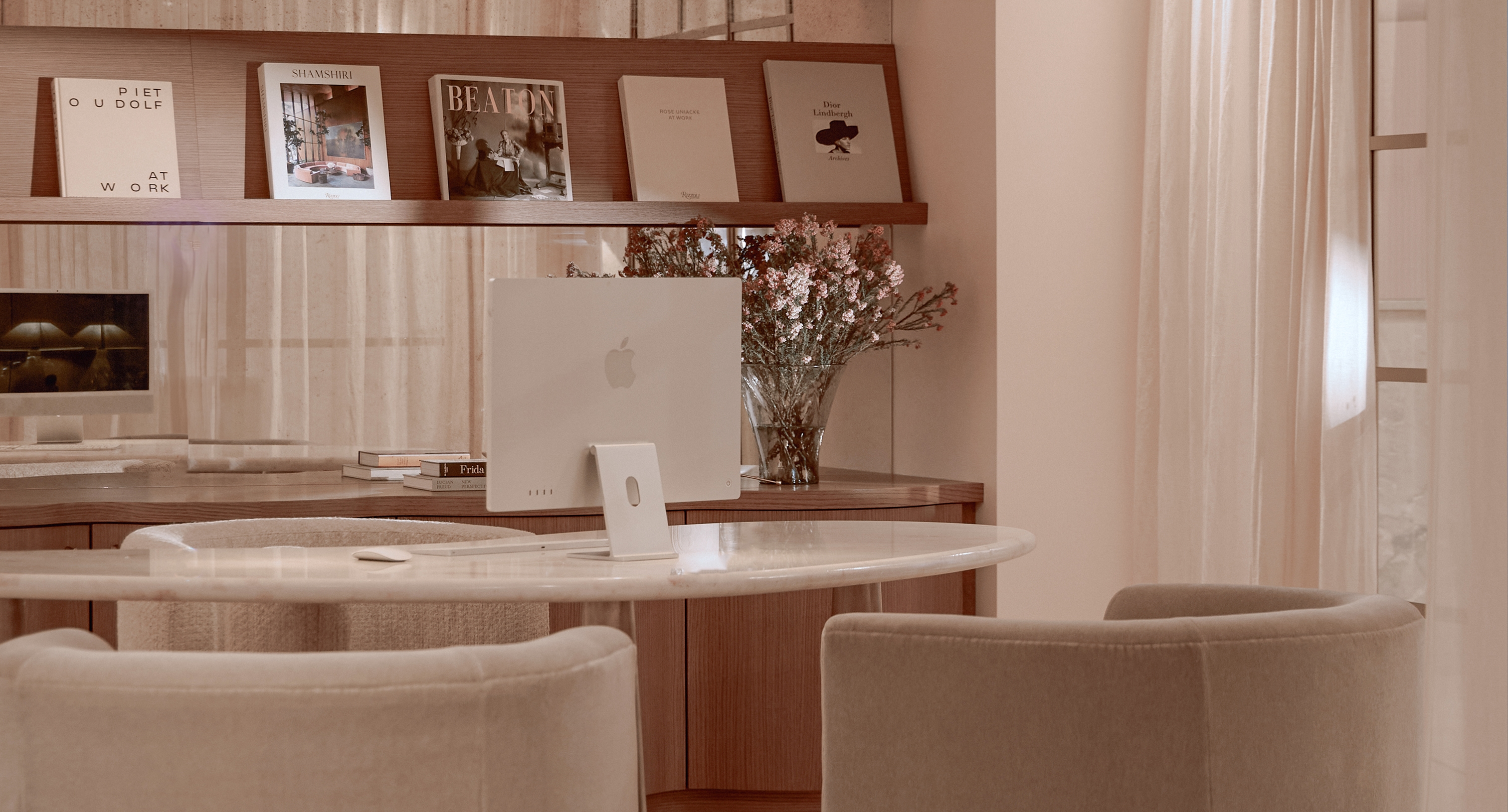 Round marble desk with upholstered chairs, computer workstation, books, and floral arrangement in a softly lit interior